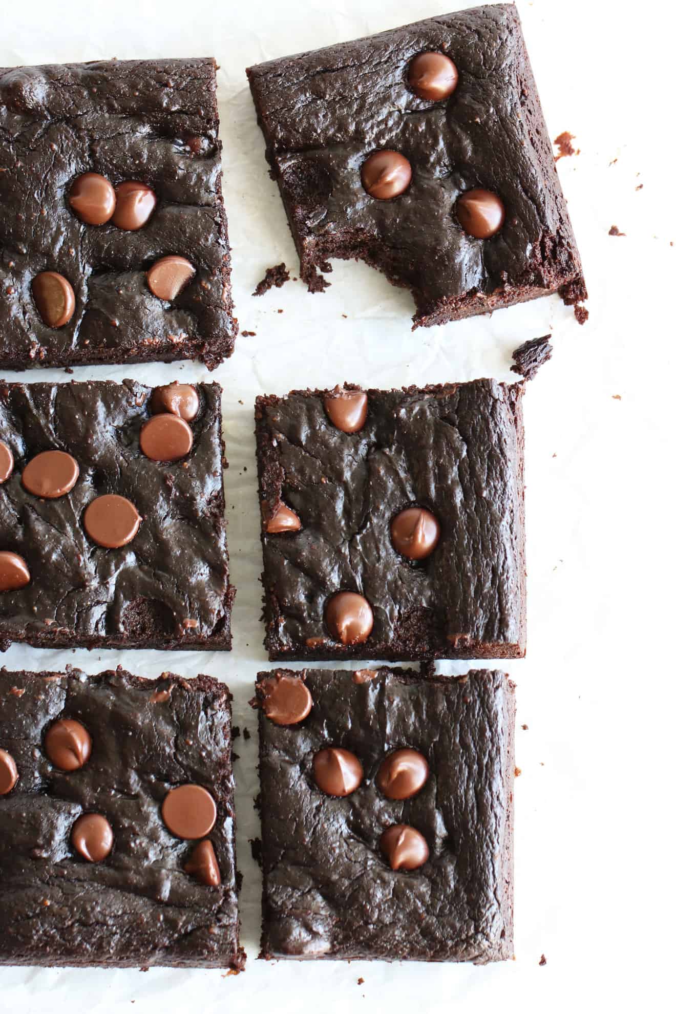 This is an overhead image of healthy avocado brownies with chocolate chips on the tops. The brownies are on a white surface and one brownie square has a bite taken out of it. 