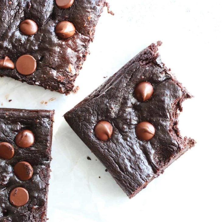 This is an overhead image of dark chocolate brownies with chocolate chips on the tops. The brownies are on a white surface and one brownie square has a bite taken out of it.