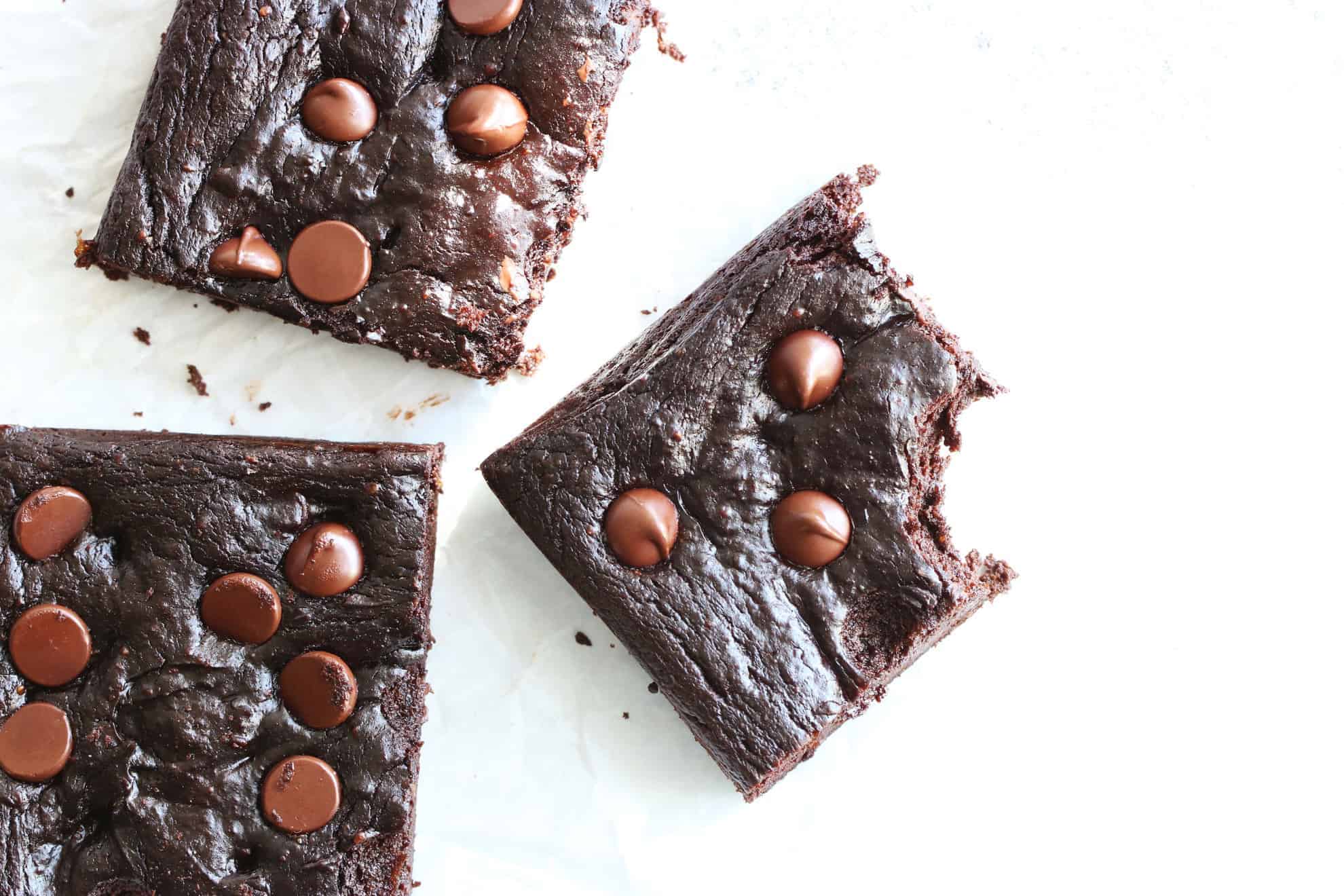 This is an overhead image of dark chocolate brownies with chocolate chips on the tops. The brownies are on a white surface and one brownie square has a bite taken out of it. 