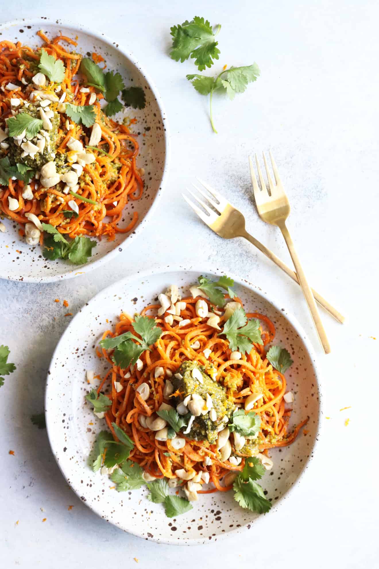This is an overhead image of a speckled bowl with sweet potato noodles, pesto, cilantro, and cashews. The bowl sits on a white surface and another bowl to the top left of the image. Gold forks lay on the surface to the top of the bowl.