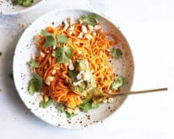 This is an overhead image of a speckled bowl with sweet potato noodles, pesto, cilantro, and cashews. The bowl sits on a white surface and another bowl to the top left of the image. A gold fork is in the bowl twirling the noodles.