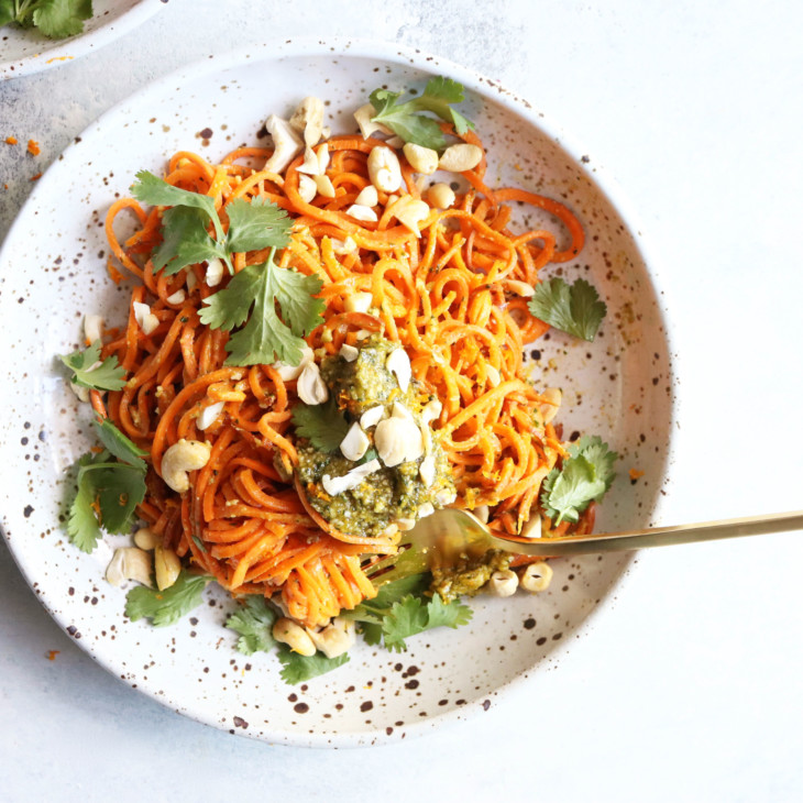 This is an overhead image of a speckled bowl with sweet potato noodles, pesto, cilantro, and cashews. The bowl sits on a white surface and another bowl to the top left of the image. A gold fork is in the bowl twirling the noodles.