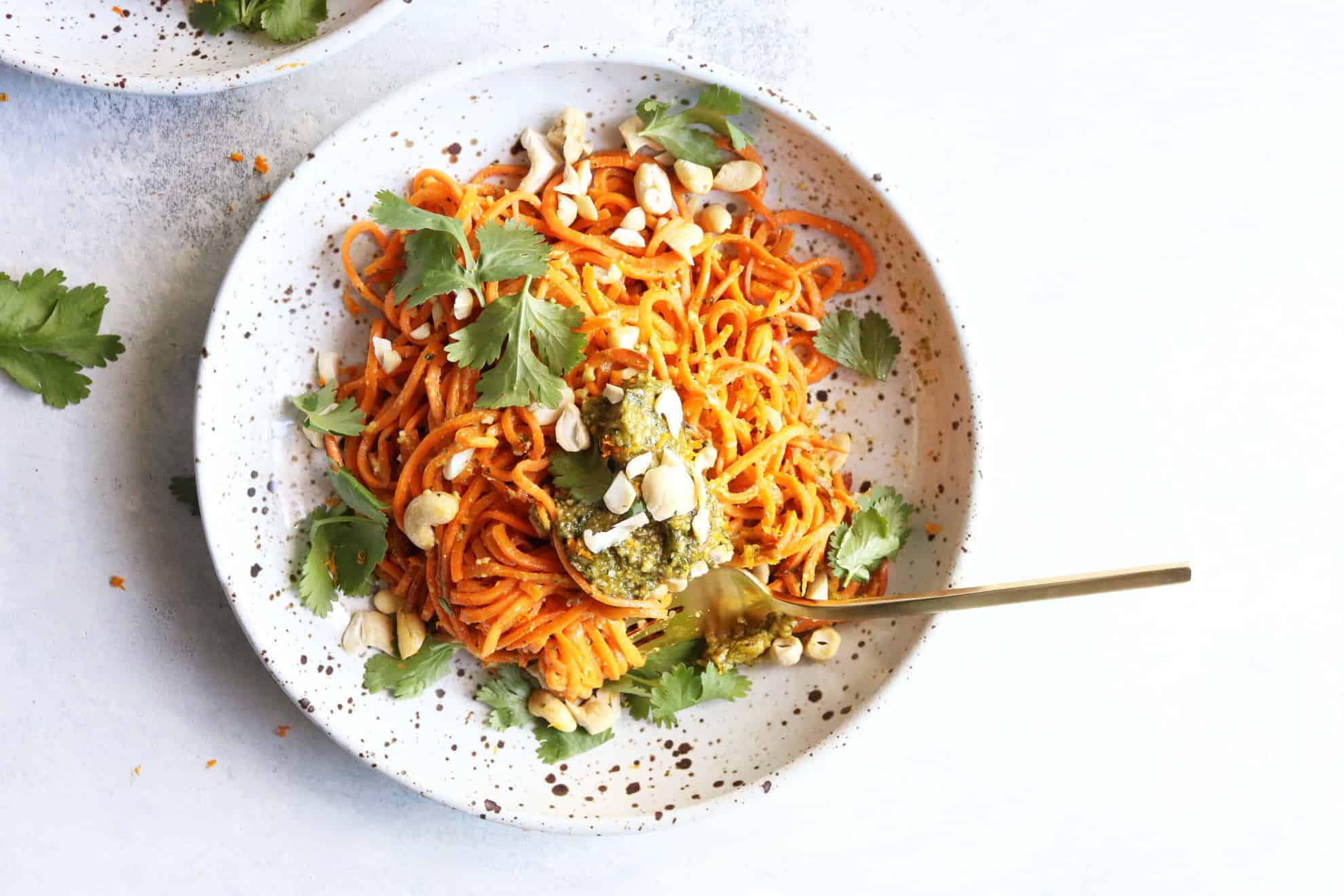 This is an overhead image of a speckled bowl with sweet potato noodles, pesto, cilantro, and cashews. The bowl sits on a white surface and another bowl to the top left of the image. A gold fork is in the bowl twirling the noodles.