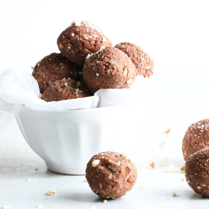 small white bowl filled with chocolate brazil nut energy balls with three energy balls sitting on the white counter