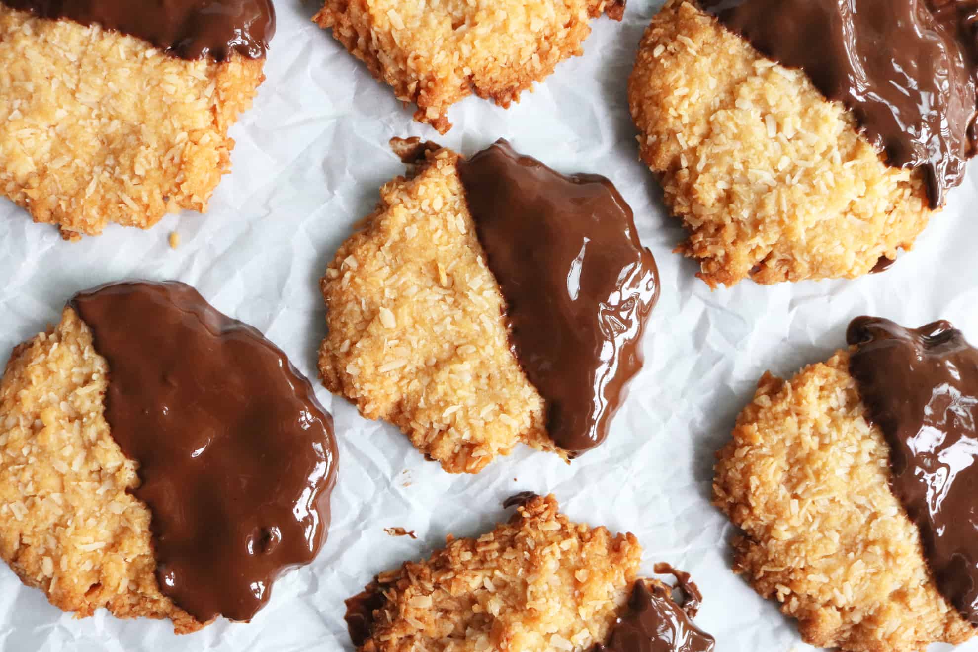 This is an overhead image of coconut cookies dipped half in melted chocolate. The cookies are on a white piece of parchment paper. 