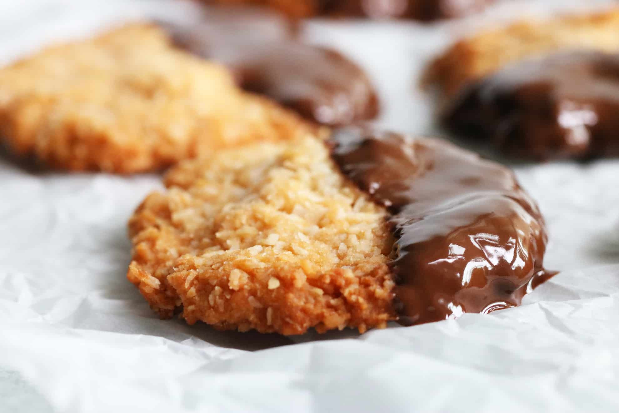 This is a side view of a coconut cookie dipped in melted chocolate. The cookie sits on a white piece of parchment paper with more cookies blurred in the background. 