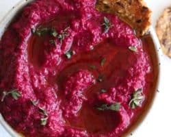 This is an overhead image of a speckled white bowl on a white counter. Inside the bowl is a pink beet dip topped with oil and fresh thyme leaves. Crackers are off to the side, next to the bowl on the white counter.
