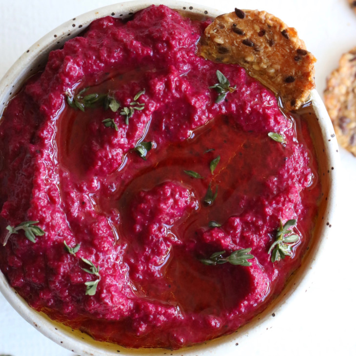 This is an overhead image of a speckled white bowl on a white counter. Inside the bowl is a pink beet dip topped with oil and fresh thyme leaves. Crackers are off to the side, next to the bowl on the white counter.