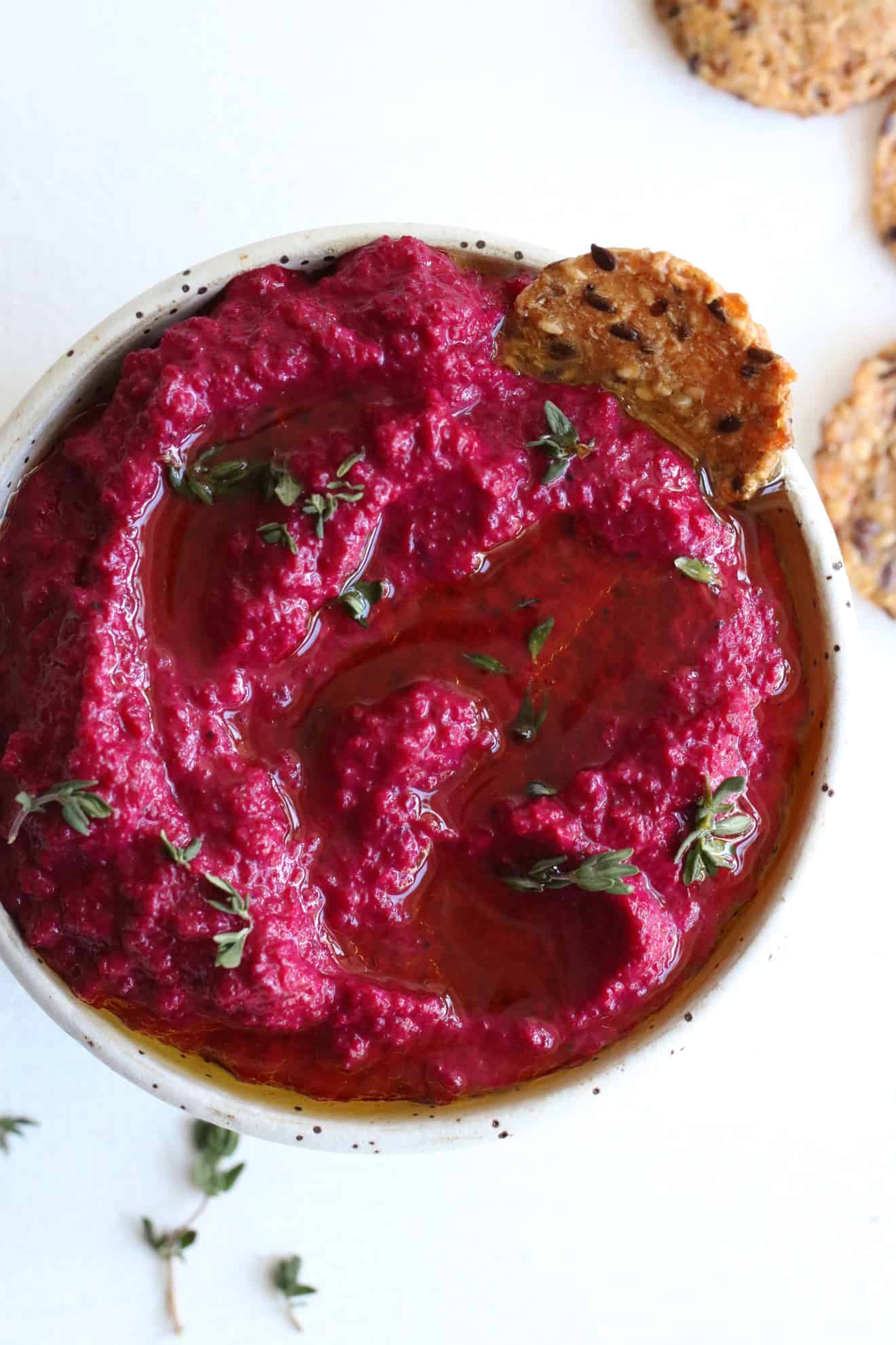 This is an overhead image of a speckled white bowl on a white counter. Inside the bowl is a pink beet dip topped with oil and fresh thyme leaves. Crackers are off to the side, next to the bowl on the white counter. One cracker is dipping into the dip and leaning against the side of the bowl. 