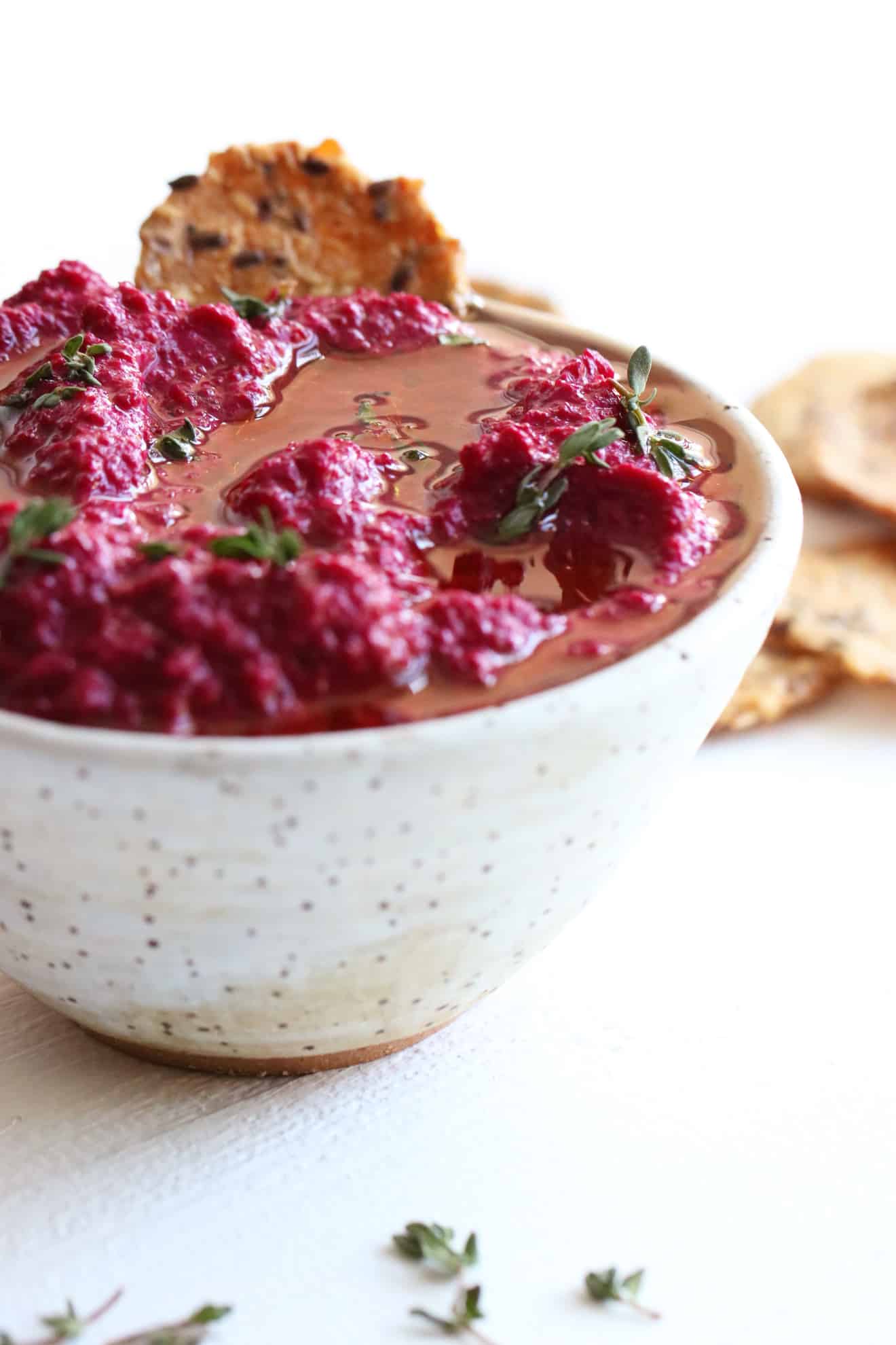 This is a side view of a speckled white bowl on a white counter. Inside the bowl is a pink beet dip topped with oil and fresh thyme leaves. Crackers are off to the side, next to the bowl on the white counter.