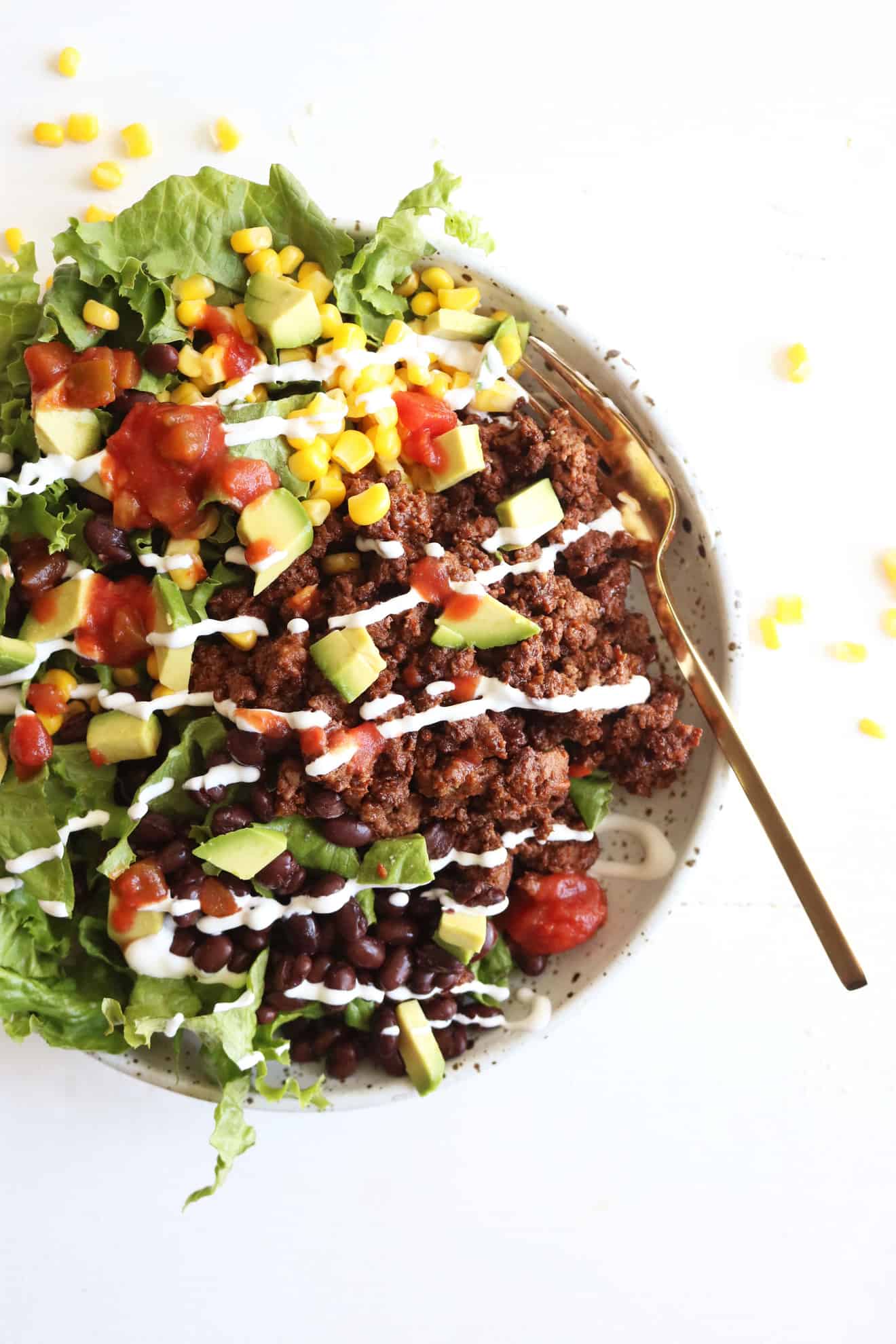 This is an overhead image of a bowl filled with lettuce, ground beef, salsa, avocado, corn, black beans, and sour cream. The bowl sits on a white counter.