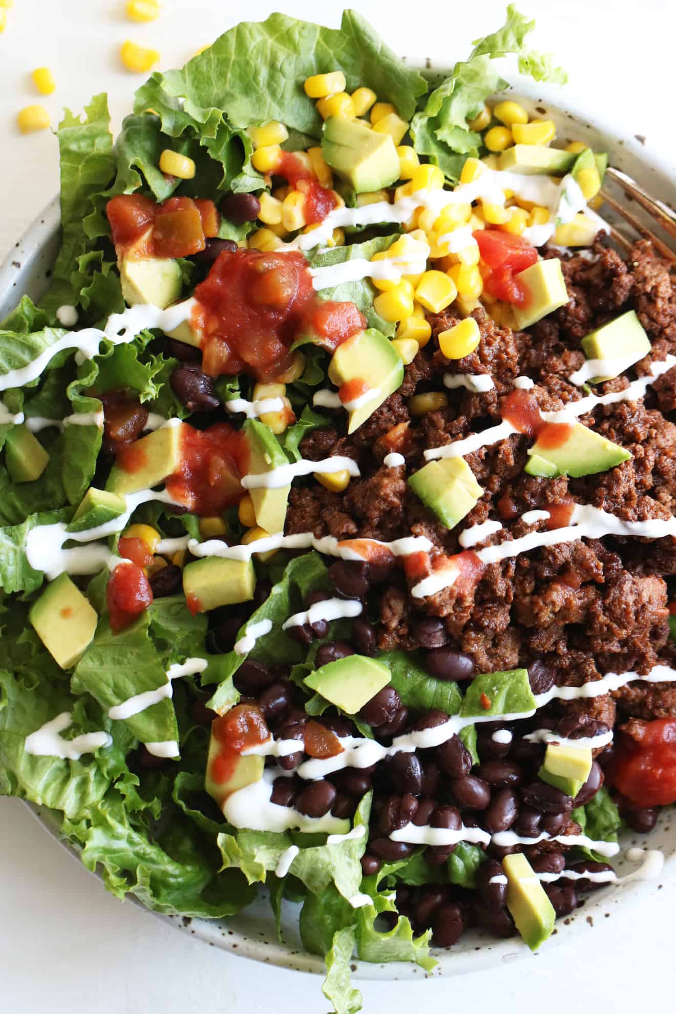 This is an overhead image of a bowl filled with lettuce, ground beef, salsa, avocado, corn, black beans, and sour cream. The bowl sits on a white counter and a gold fork lies partially in the bowl. 