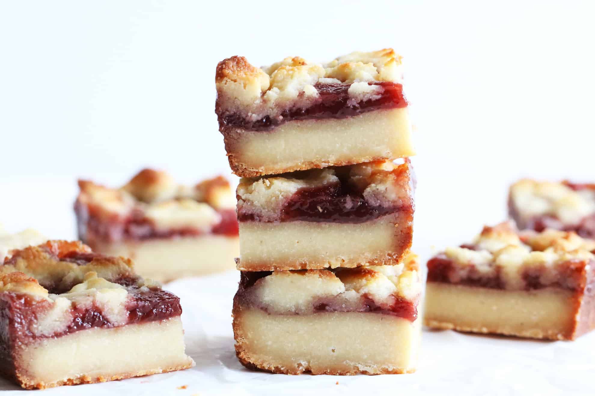 This is a side view of a stack of three Strawberry Crumble Bars. The stack sits on a white counter with more bars blurred to the side and in the background.