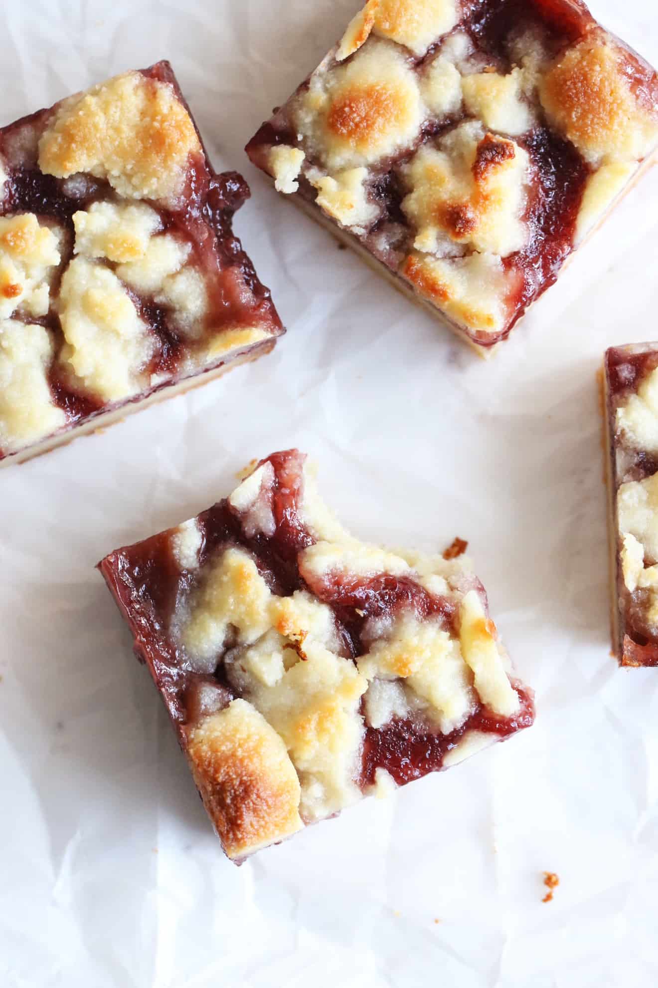 This is an overhead image of strawberry crumble bars. The bars sit on a white piece of parchment paper and one has a bite taken out of it.