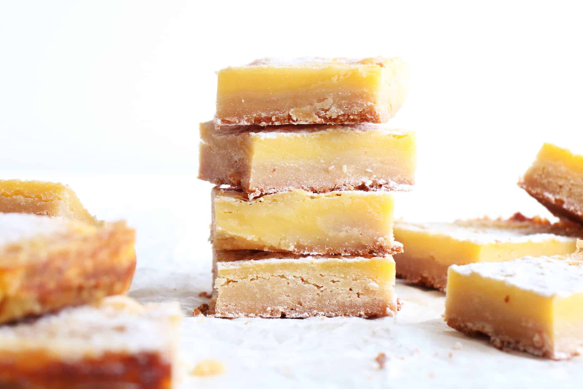 This is a side view of a stack of four lemon bars. The bars are sitting on a white piece of parchment paper with more bars around the stack.
