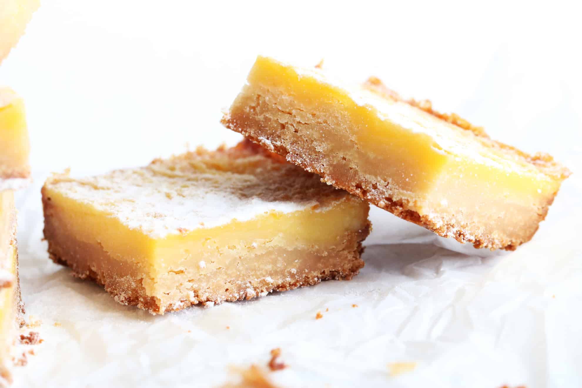 This is a side view of a lemon bar leaning against another lemon bar. The bars sit on a white piece of parchment paper with a white background. 