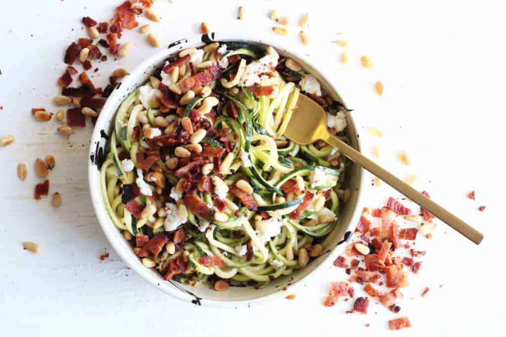 This is an overhead image of zoodles with crumbled bacon, cheese, and pine nuts. A fork twirls the zoodles and lays on the side of the bowl. 