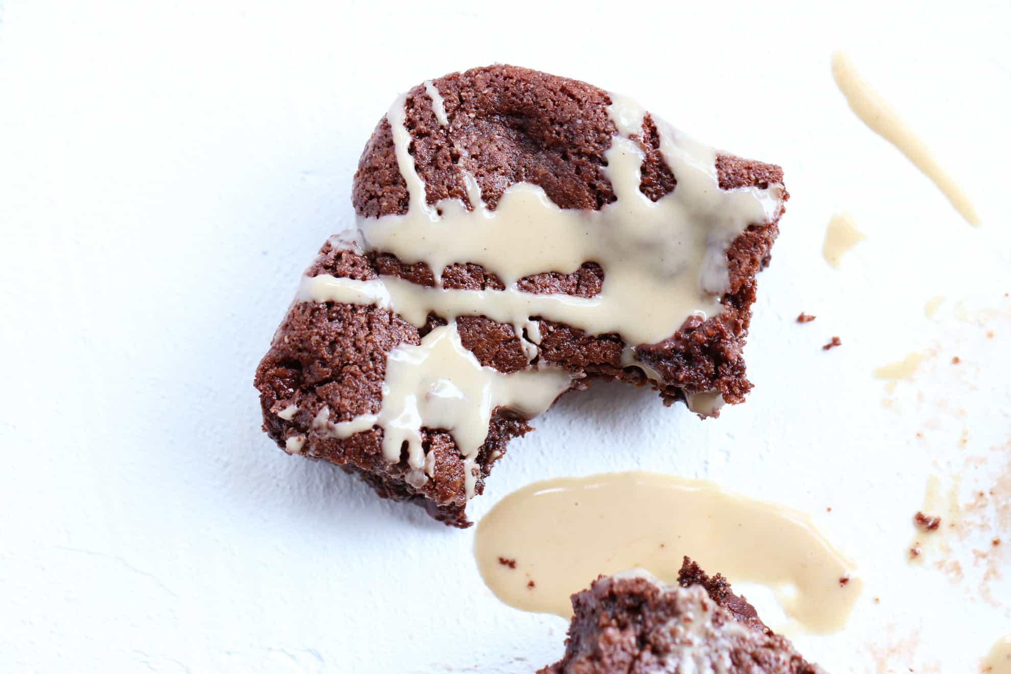 This is an overhead image of a tahini brownie with a bite taken out of it. The brownie sits on a white surface. Tahini is drizzled on the brownie and on some of the counter.