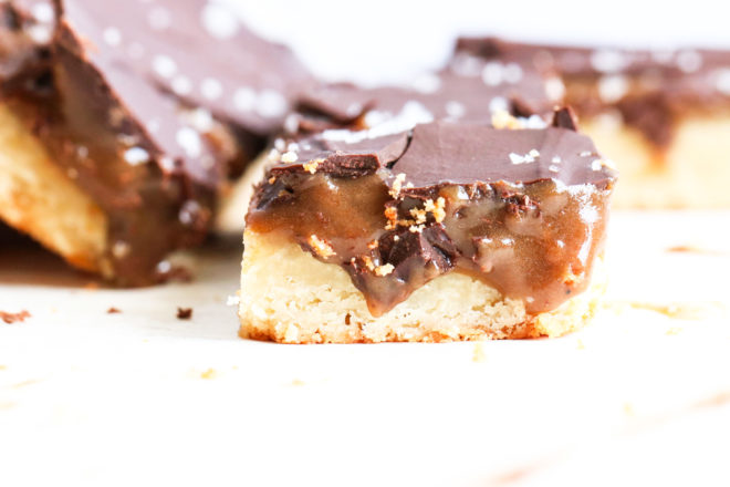 This is a side view of a homemade twix bar. The bar has three layers: shortbread, caramel, and chocolate. More twix bars are blurred in the background.