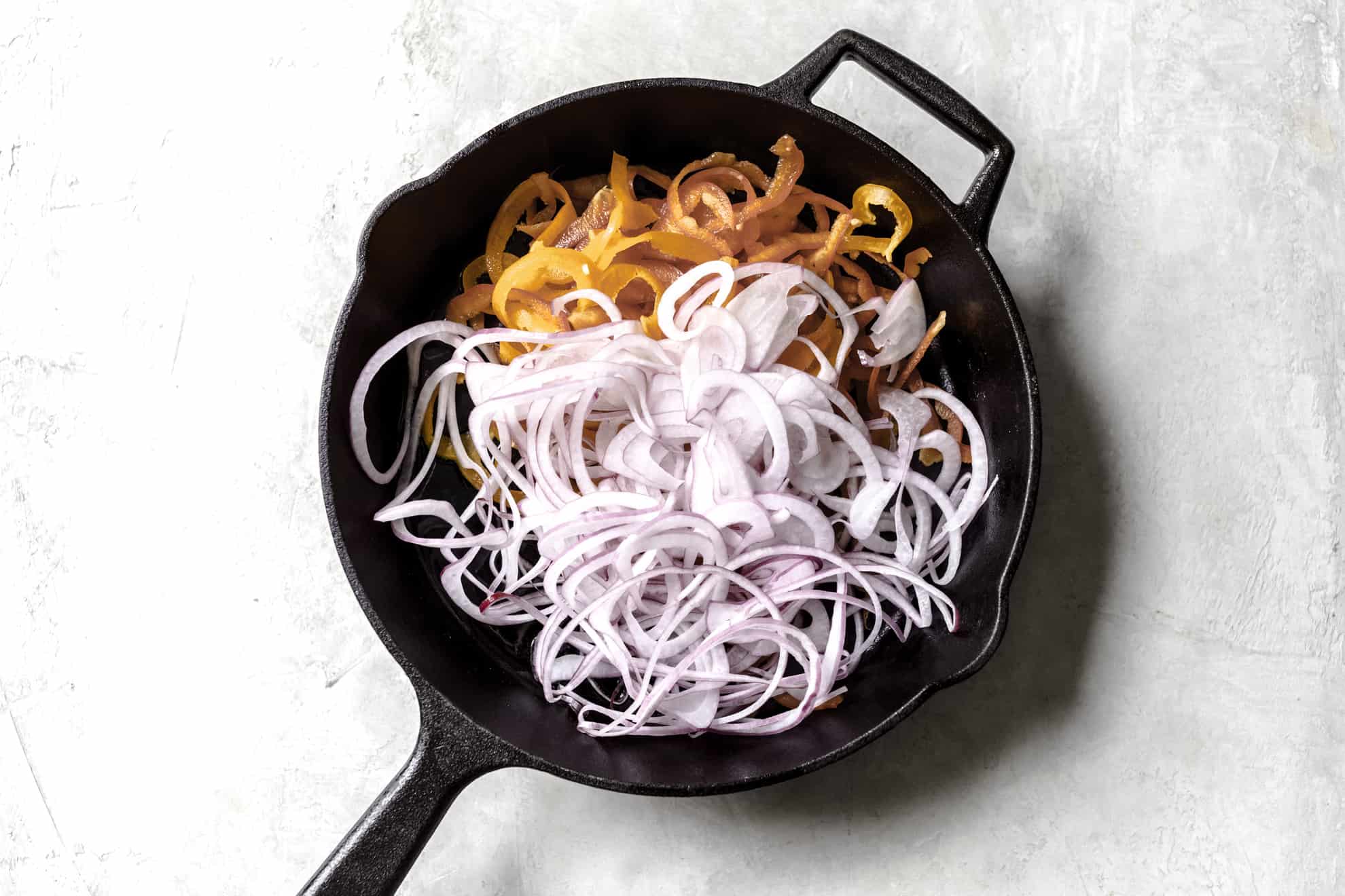 This is an overhead image of a cast iron skillet on a white surface. Inside the skillet is spiralized peppers and spiralized onions. 