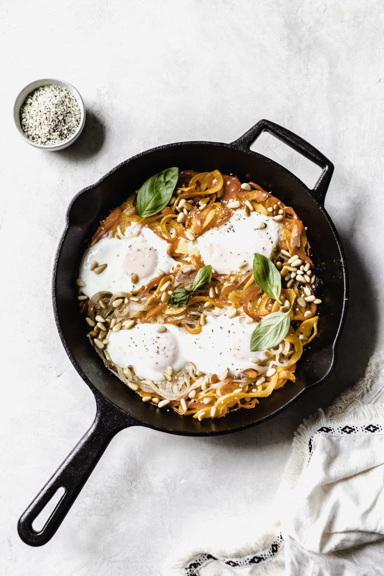 This is an overhead image of a skillet on a white surface. Inside the skillet is spiralized peppers, spiralized onions, eggs, pine nuts, and basil. A small bowl of hemp hearts is in the top left of the image. A white tea towel is in the bottom right corner of the image. 