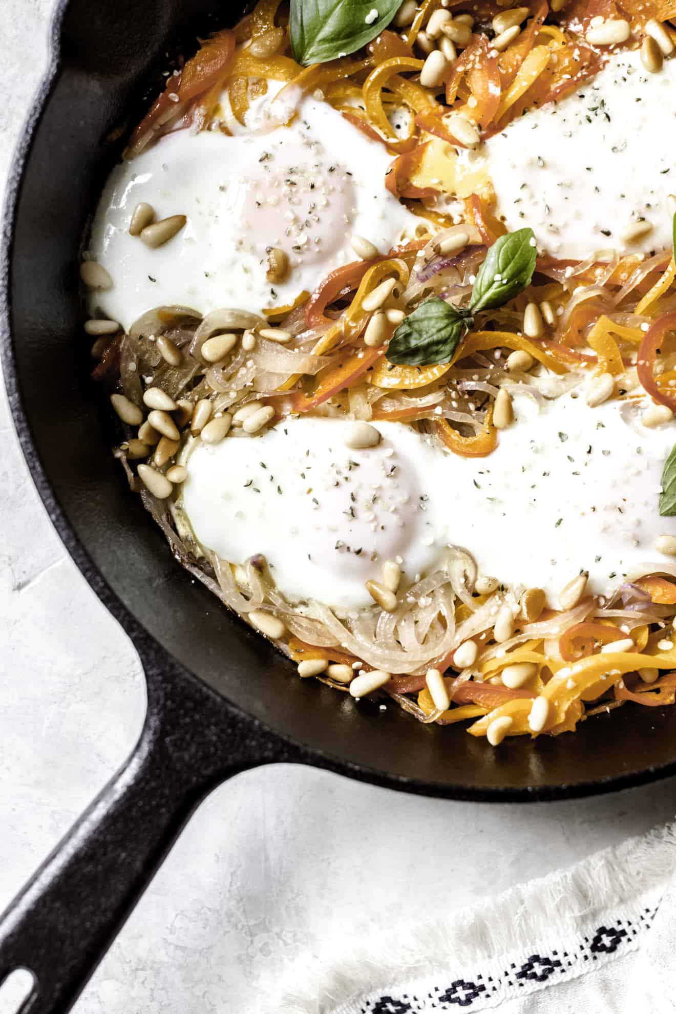 This is an overhead image of a skillet on a white surface. Inside the skillet is spiralized peppers, spiralized onions, eggs, pine nuts, and basil. A white tea towel is in the bottom right corner of the image. 