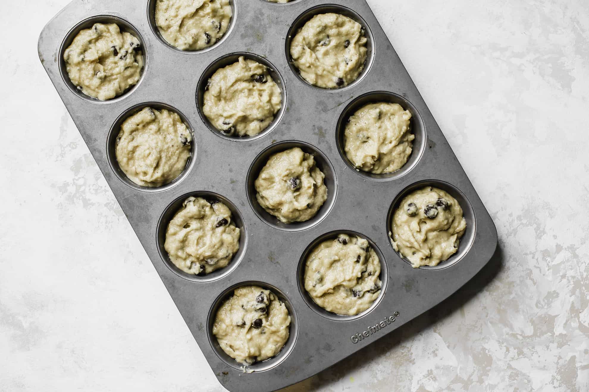 This is an overhead image of a muffin tin with raw batter inside. The muffin tin sits on a white and grey surface. Inside the tin is a raw, tan batter with some chocolate chops visible. 