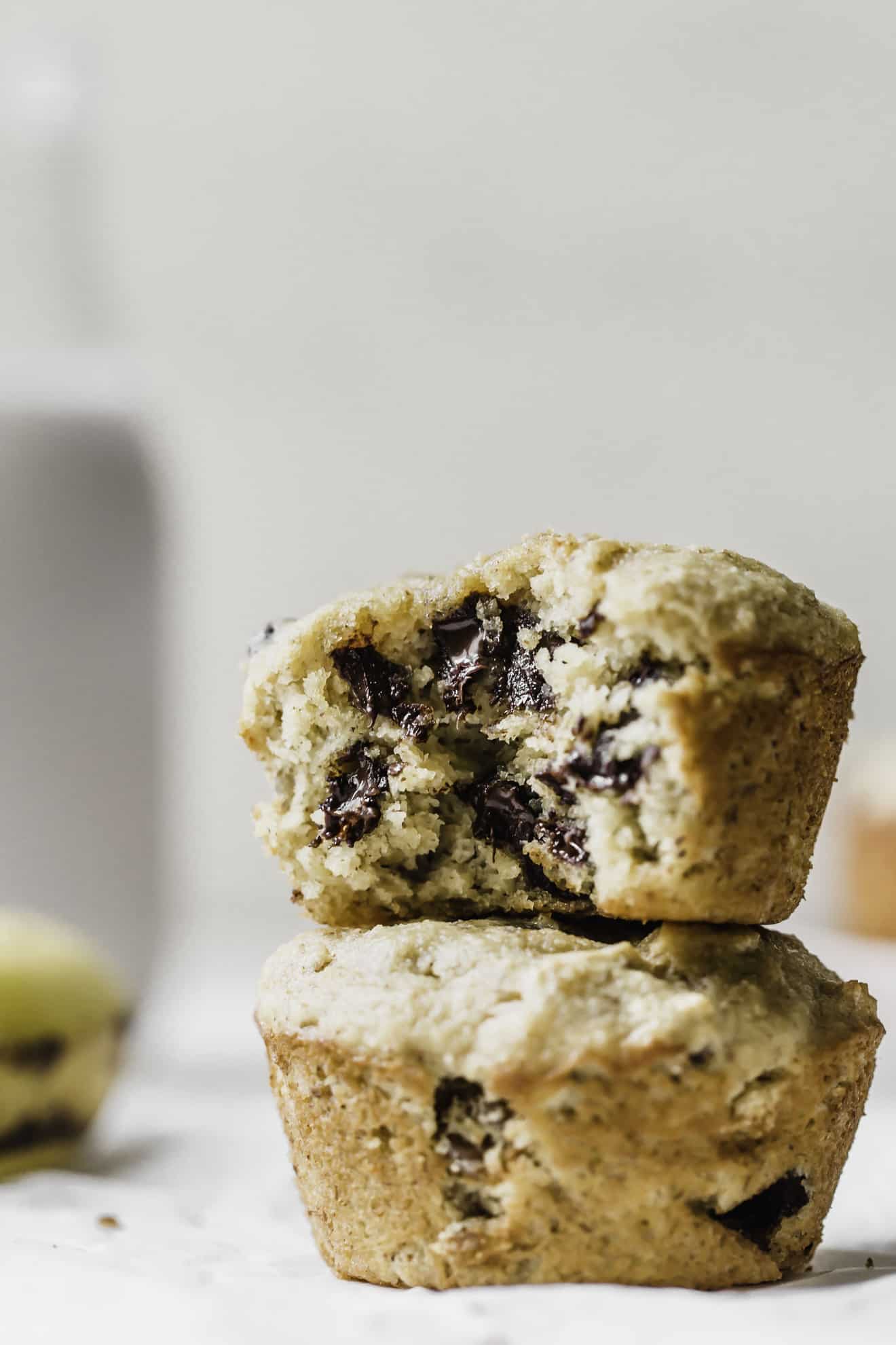 This is a side view of two banana muffins stacked on top of each other. The top muffin has a bite taken out revealing melty chocolate chips. The muffins sit on a white surface with a banana blurred in the background. 