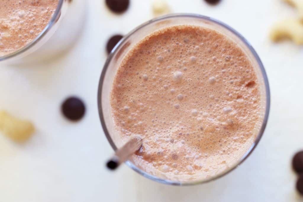 This is an overhead view of chocolate milk in a glass cup. The chocolate milk has a bot of bubbles and a stainless steep straw. Another glass is in the top left corner of the image. Cashews and chocolate chips are scattered around the white counter. 