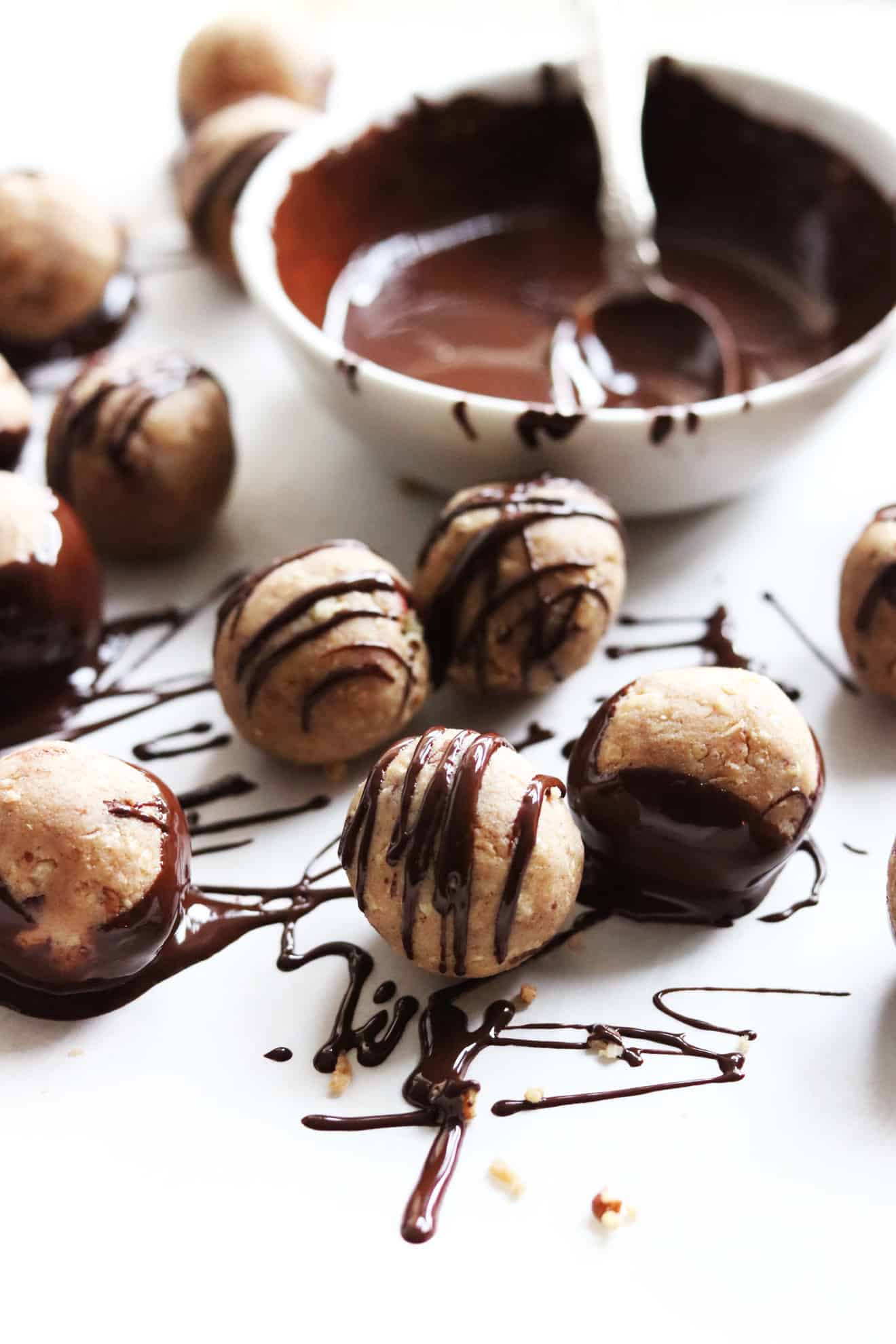 Side view of gingerbread snack balls on a white counter with melted chocolate drizzled on top and a small bowl with melted chocolate and a spoon blurred in the background