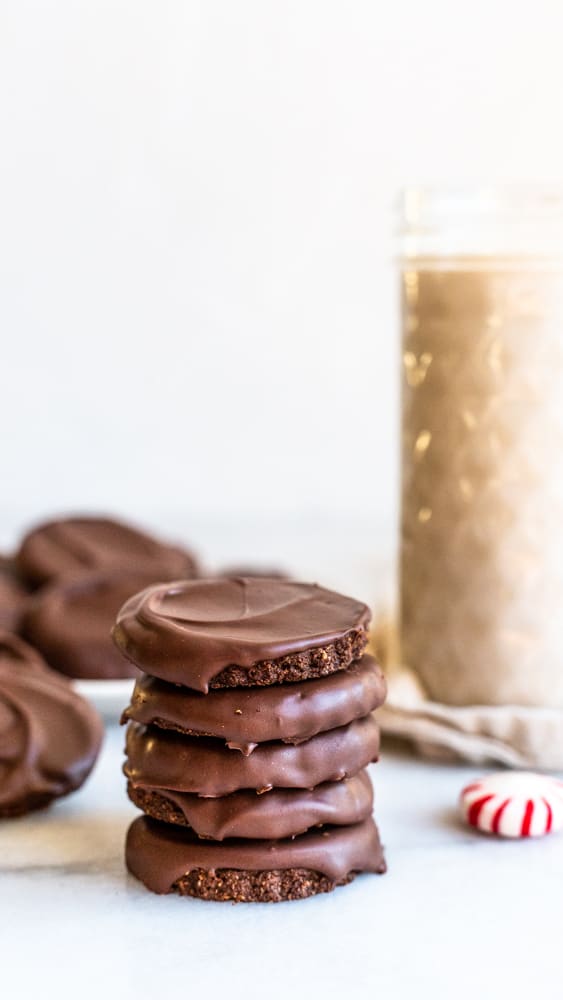 This is a side view of a stack of think mint cookies. The stack sits on a white counter with more chocolate thin mint cookies, a glass of light brown liquid, and some peppermint candies blurred in the background.