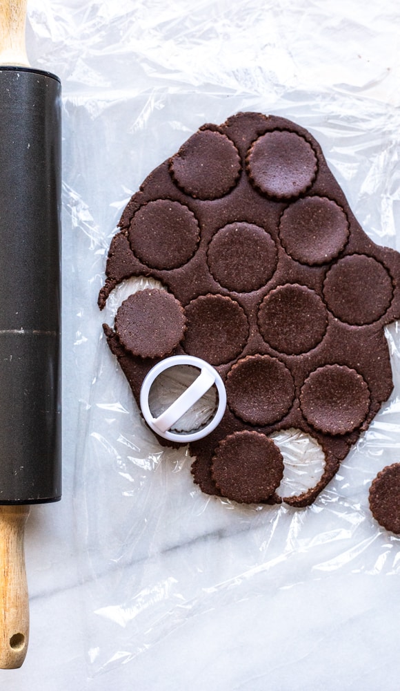 This is an overhead image of chocolate cookie dough rolled out on plastic wrap. The dough is cut with circle cookie cutters. A rolling pin is to the left of the image. 