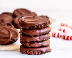 This is a side view of a stack of think mint cookies. The stack sits on a white counter with more chocolate thin mint cookies and some peppermint candies blurred in the background.