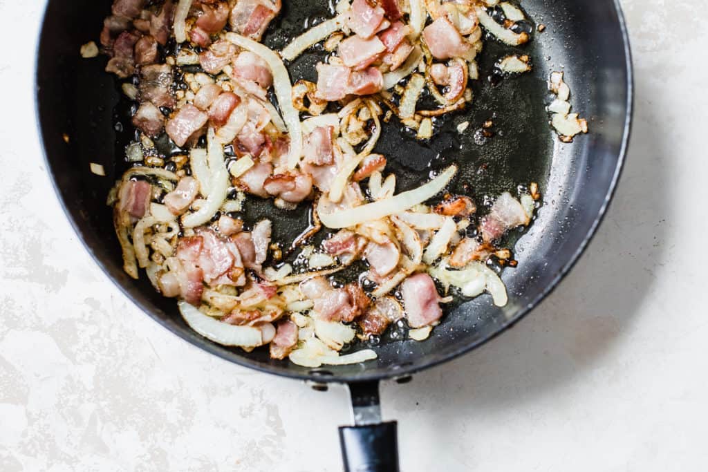 This is an overhead image of a pan with sautéed onions and bacon. The pan sits on a white and tan background. 