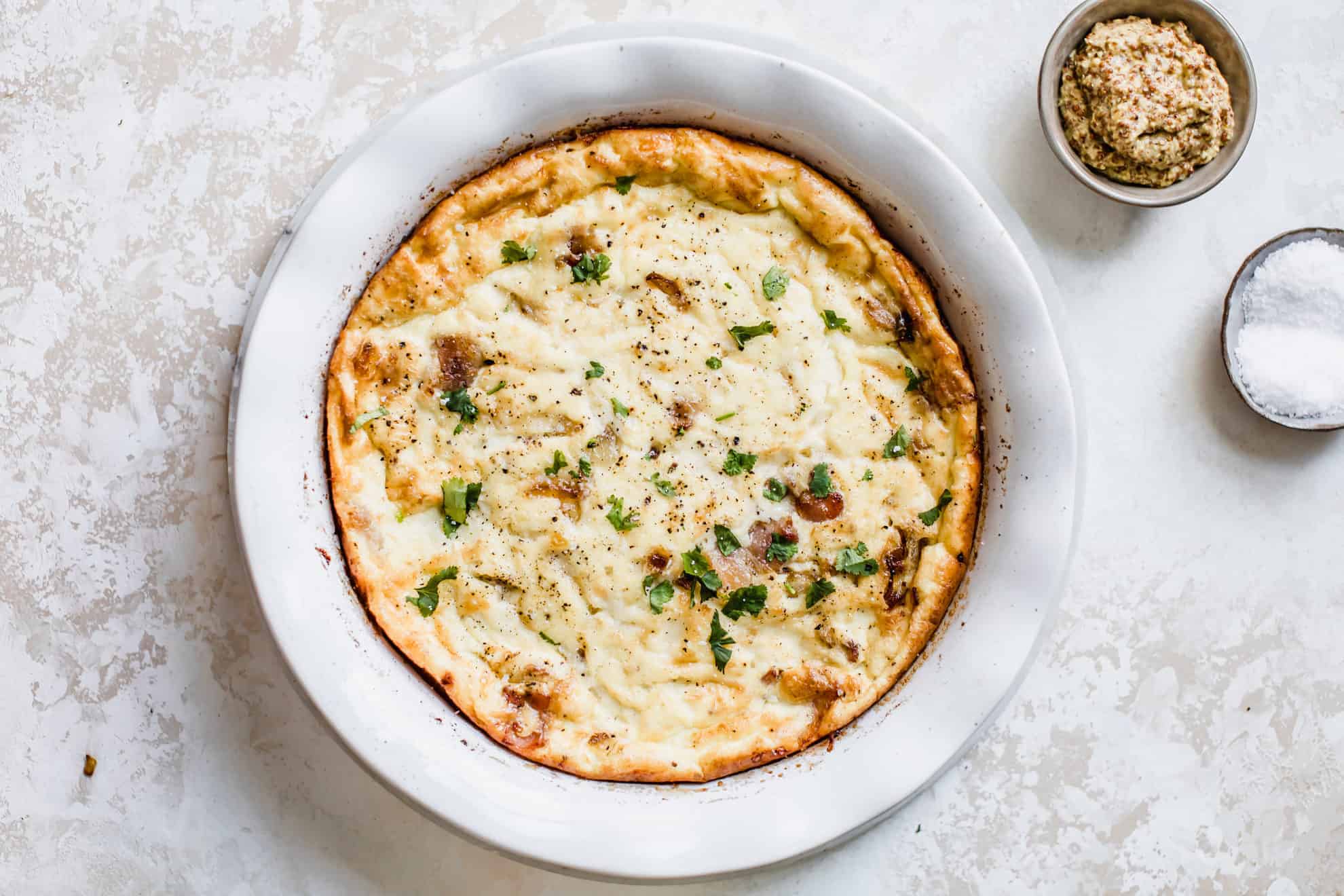 This is an overhead image of a baked crustless quiche dish on a white counter. The quiche has bits of chopped herbs and small bowls of mustard and salt to the side. 