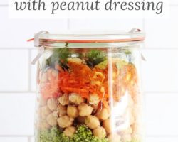 This is a side view of a glass jar with a lid. Inside the jar is layers of broccoli, chickpeas, carrots, and herbs. Text overlay reads "broccoli chickpea salad with peanut dressing"