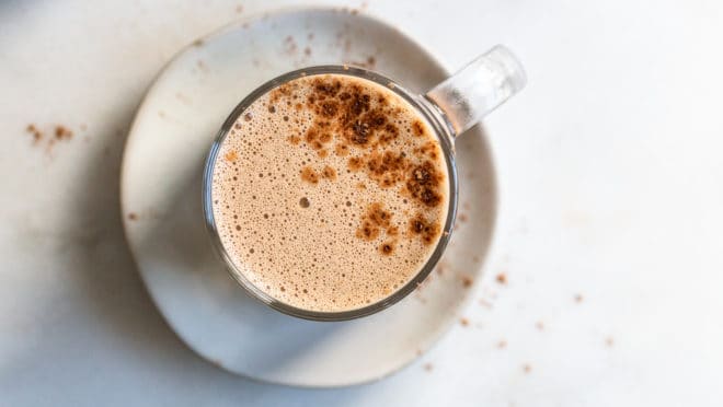 This is an overhead image of a glass mug on a white saucer sitting on a white counter. The mug has a frothy mocha in it with a sprinkle of cocoa powder. 