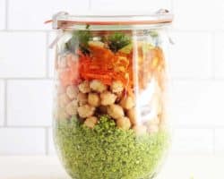 This is a side view of a glass jar with a lid. Inside the jar has broccoli rice at the bottom, chickpeas, carrots, and fresh herbs.