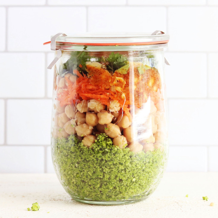 This is a side view of a glass jar with a lid. Inside the jar has broccoli rice at the bottom, chickpeas, carrots, and fresh herbs.