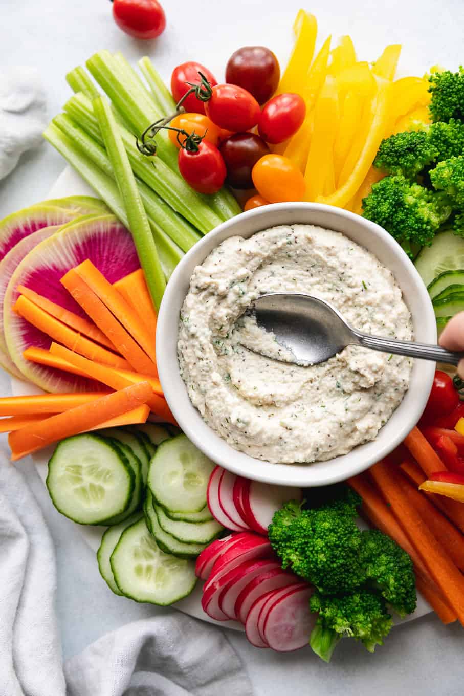 This is an overhead image of a white bowl filled with a creamy dip. Around the dip are fresh veggies like cut peppers, radish, carrots, cucumbers, and celery. A spoon is spreading the dip out in the bowl.