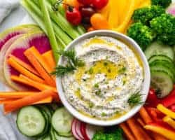 This is an overhead image of a white bowl filled with a creamy dip. The dip is topped with a drizzle of olive oil and fresh herbs. Around the dip are fresh veggies like cut peppers, radish, carrots, cucumbers, and celery.