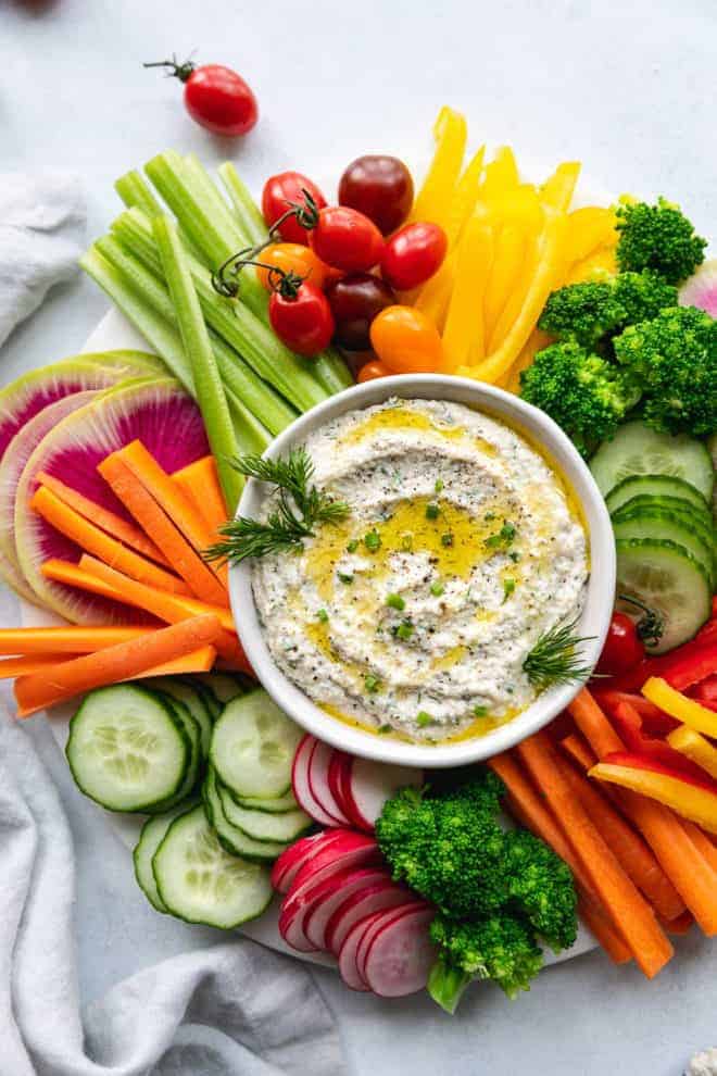 This is an overhead image of a white bowl filled with a creamy dip. The dip is topped with a drizzle of olive oil and fresh herbs. Around the dip are fresh veggies like cut peppers, radish, carrots, cucumbers, and celery.