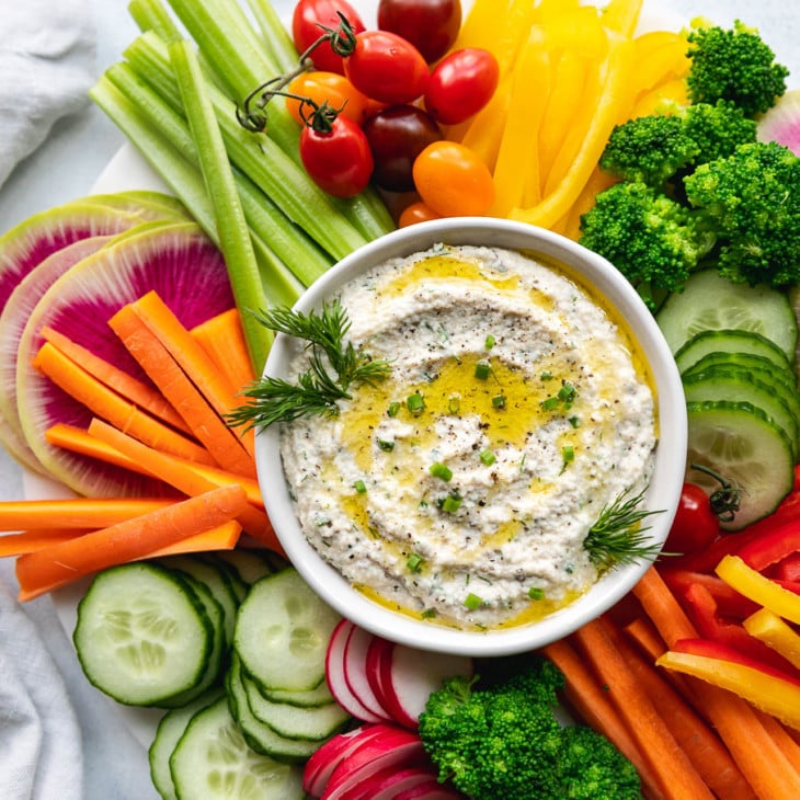 This is an overhead image of a white bowl filled with a creamy dip. The dip is topped with a drizzle of olive oil and fresh herbs. Around the dip are fresh veggies like cut peppers, radish, carrots, cucumbers, and celery.
