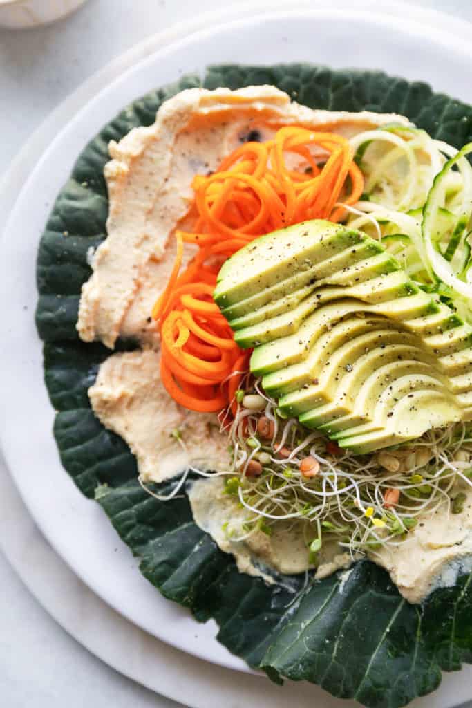 This is an overhead image of a collard green laid out on a plate. Inside the collard green leaf is hummus, spiralized veggies, sprouts and avocado. 
