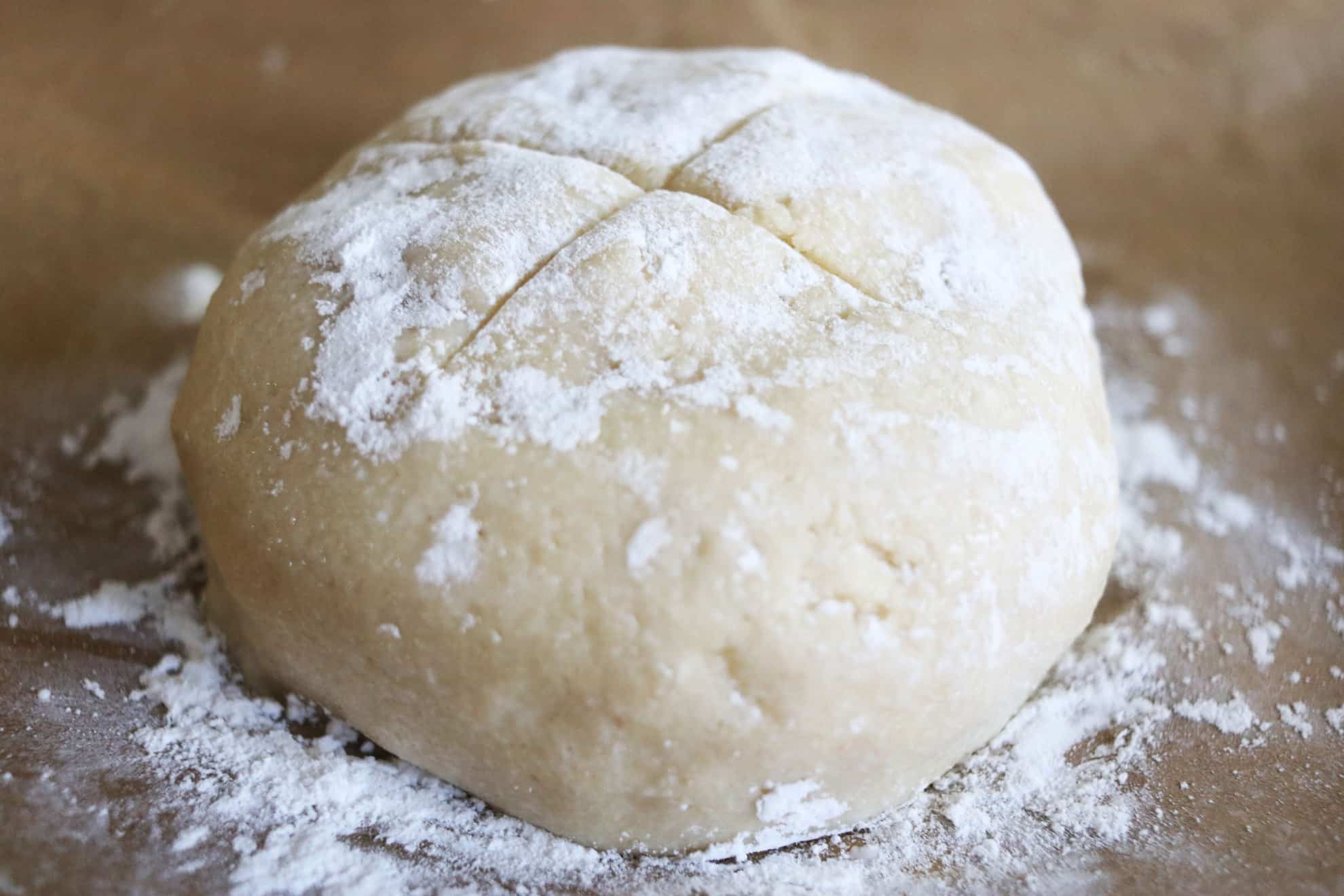 This is a side view of a ball of soda bread dough on a piece of brown parchment paper. The bread has a X in the center and is dusted with flour.