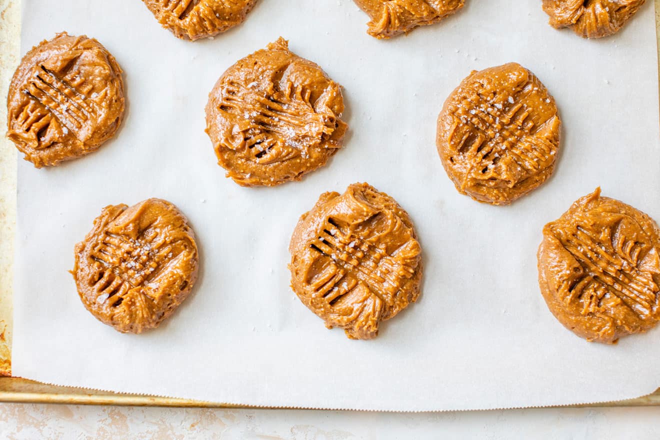 This is an overhead image of raw peanut butter cookie dough on a baking sheet lined with parchment paper. The cookies have fork imprints and salt sprinkled on top. The baking sheet is on a white and tan background. 