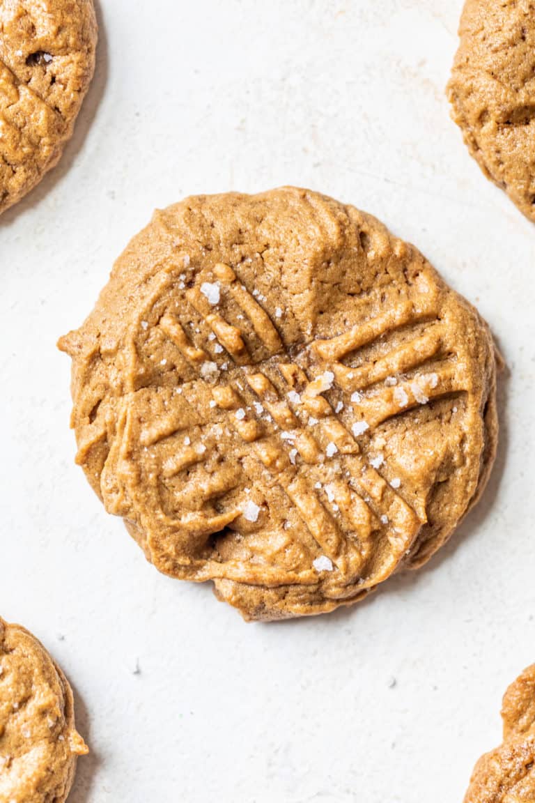 This is a close up image of a peanut butter cookie with fork imprints and salt on top. Other cookies are in the corners of the image. The cookies sit on a white background.