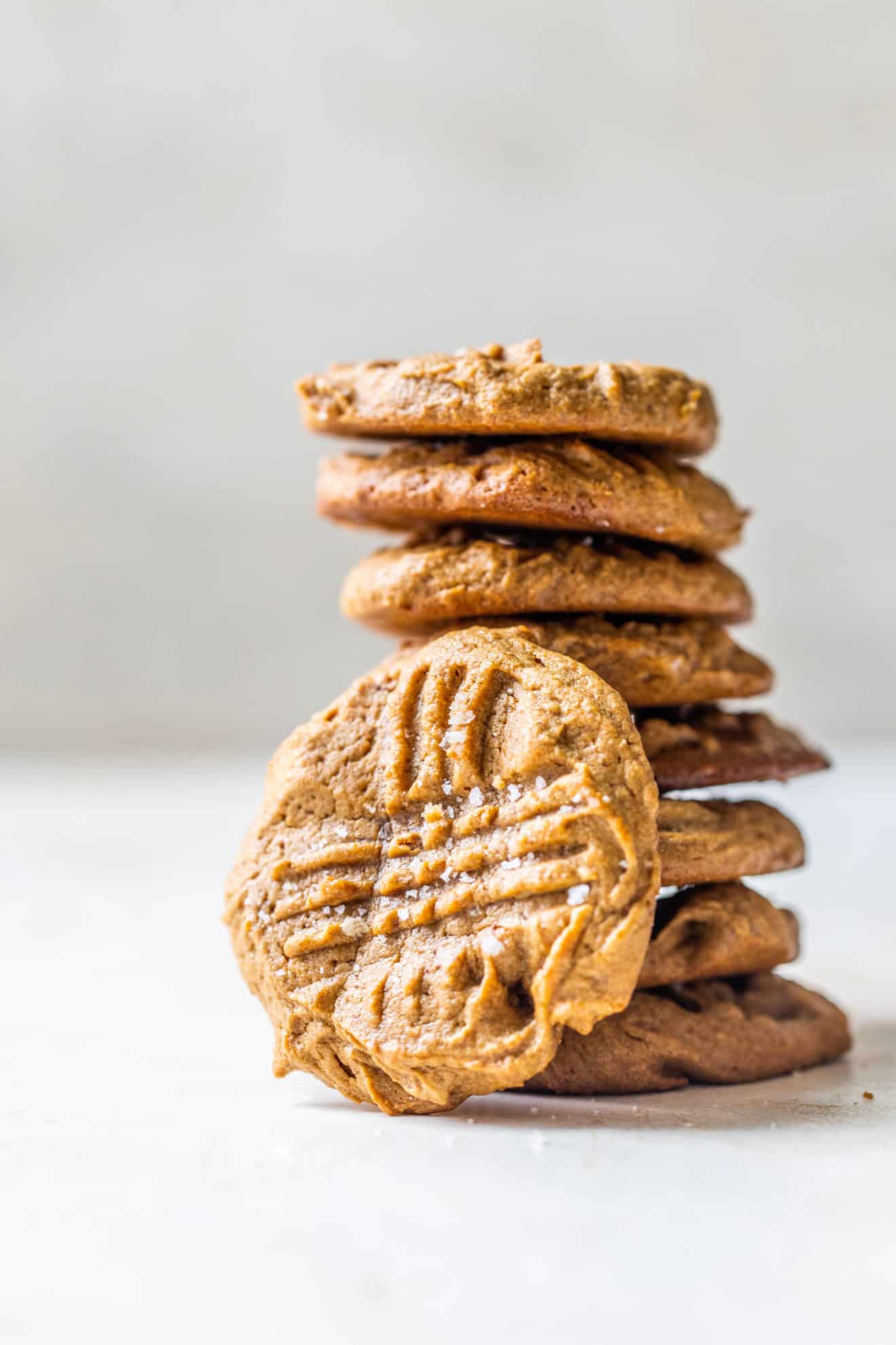 This is a side view of a stack of peanut butter cookies sitting on a white counter and a white background. One cookie leans against the stack of cookies. 