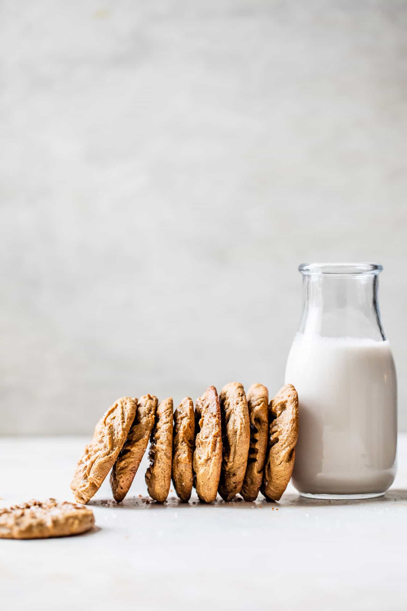 This is a side view of a a line of cookies leaning against a glass of milk. The cookies and milk are sitting on a white counter and a white background. 