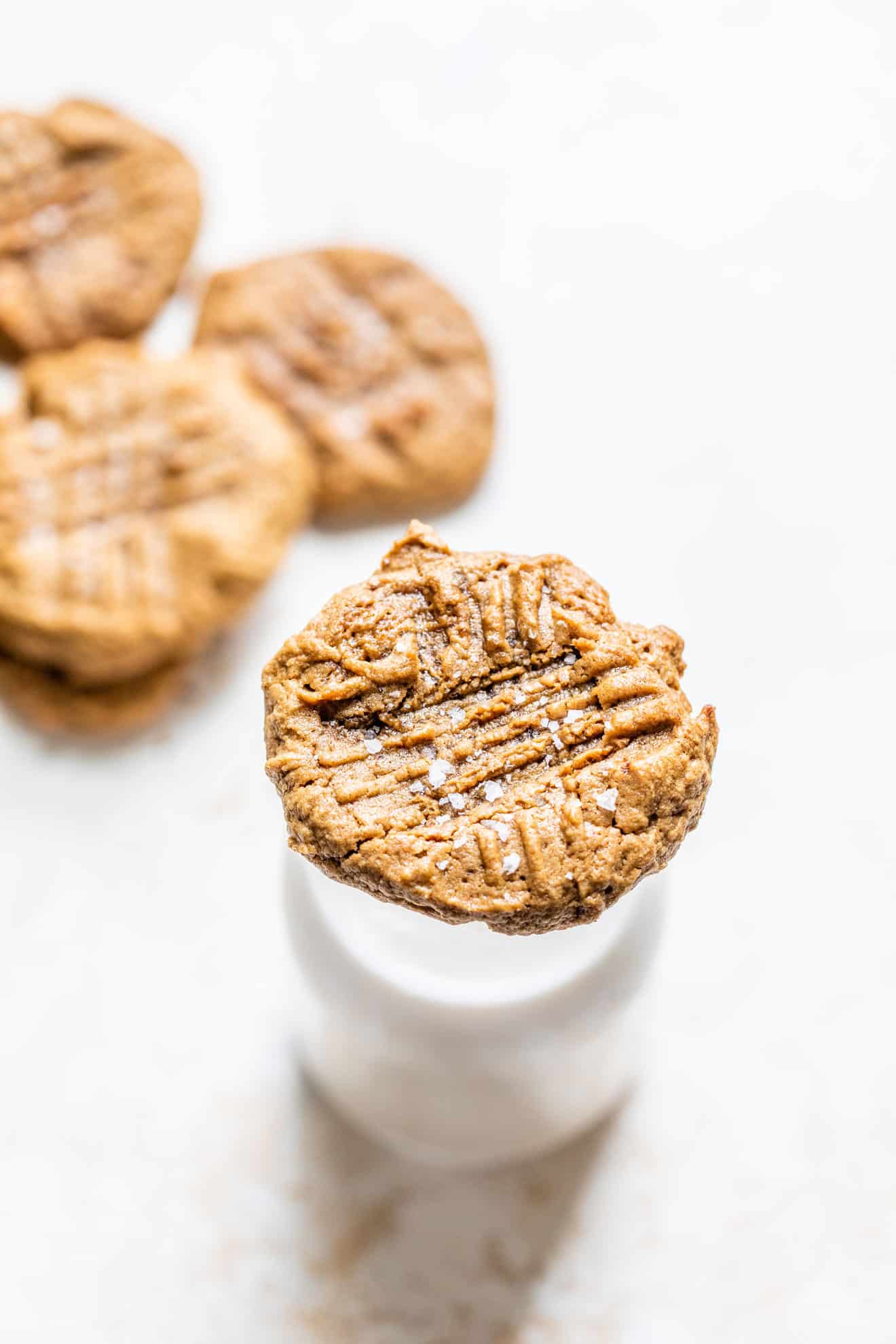 This is an overhead image of a peanut butter cookie sitting on top of a glass of milk. The milk sits on a white counter with more peanut butter cookies blurred in the background. The cookie has fork imprints and salt sprinkled on top. 